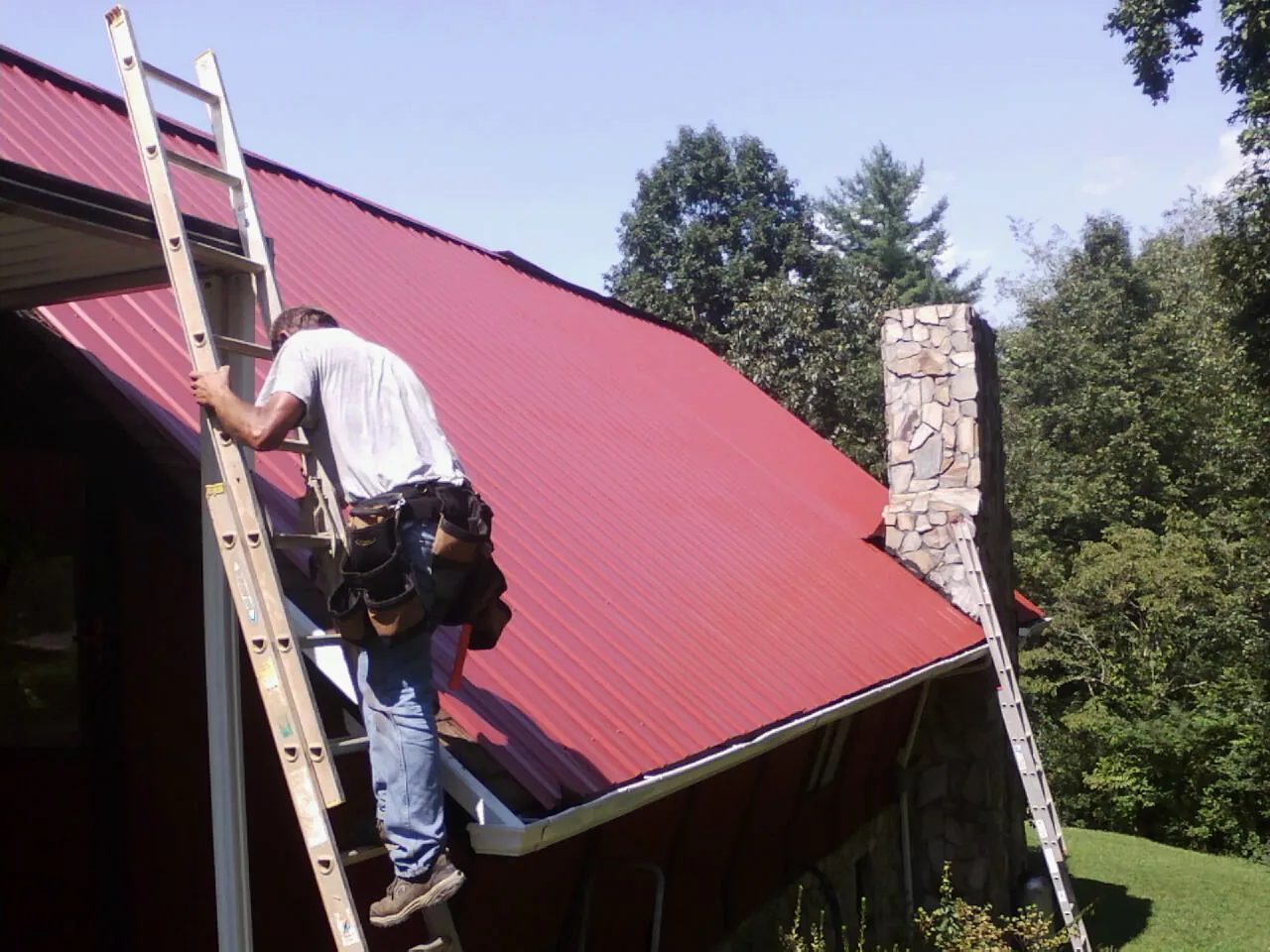 Roofer on a ladder beside a red metal roof and stone chimney — Soffit & Fascia Repair by Roofing Brothers in Jonas Ridge